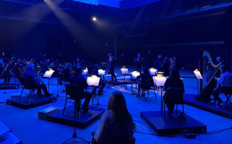 The orchestra, seated on a darkened stage that is lit from above with blue light, waits for a cue from conductor Rob Gardner.