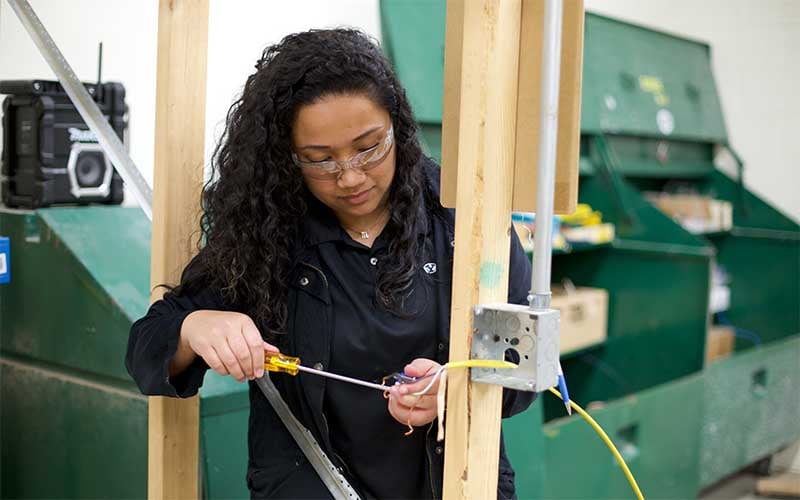 Female student with safety goggles using a screwdriver to wire a house frame.