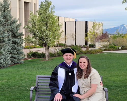 Ryan Cheney in his cap and gown after graduation sitting on a bench with his wife