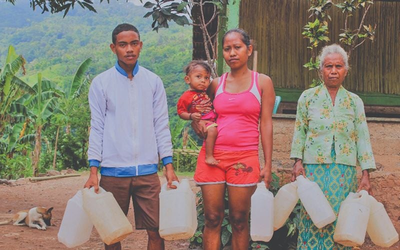 Photo of two women, a man, and a baby. They stand outside near a clean water well.