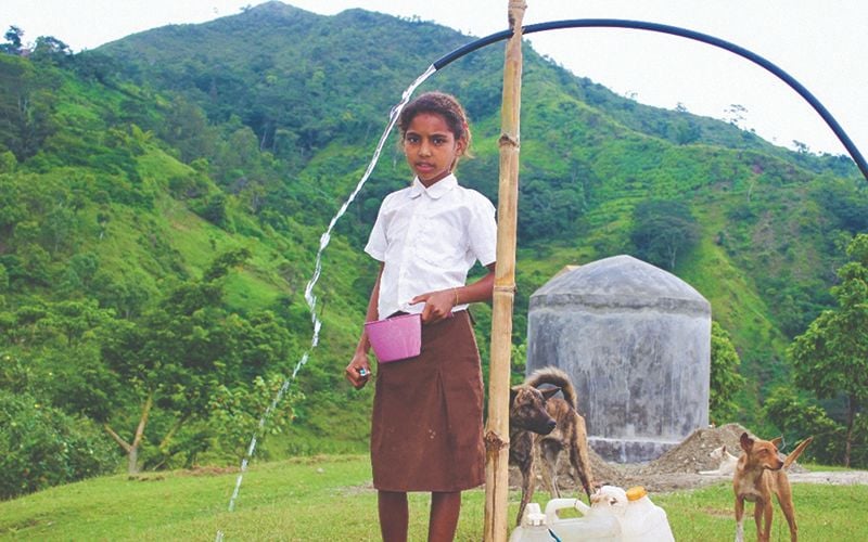 Young girl stands outside near a rubber hose filling her cup with clear running water.