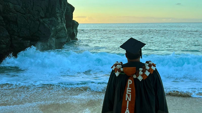 A man wearing a graduation gown standing on the beach facing the ocean.