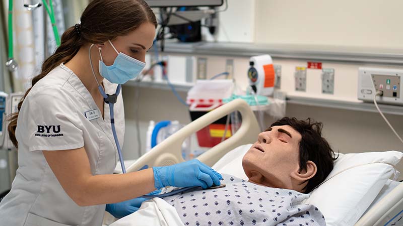 BYU Student, Camie Mendon, in nursing uniform wearing a mask and gloves, working with a medical “dummy” patient.