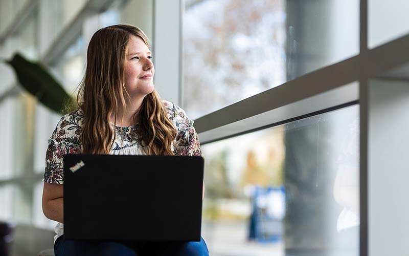 Emily Neiderhauser with her laptop by a window in the Joseph F. Smith Building.