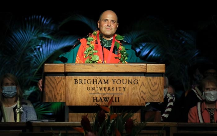 Man standing at a podium wearing academic regalia and a traditional Hawaiian cloak.