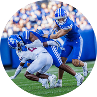 A BYU football player blocking a Southern Illinois football player while another BYU player runs the ball