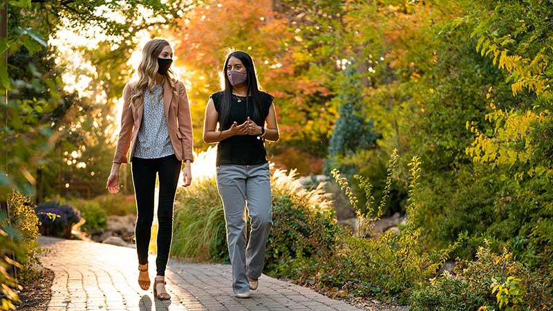Two female students wearing masks walking on campus with trees in fall colors.