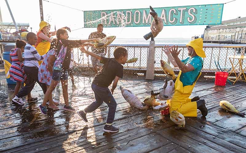 Children spray water and play with rubber fish on a waterfront with some adults.