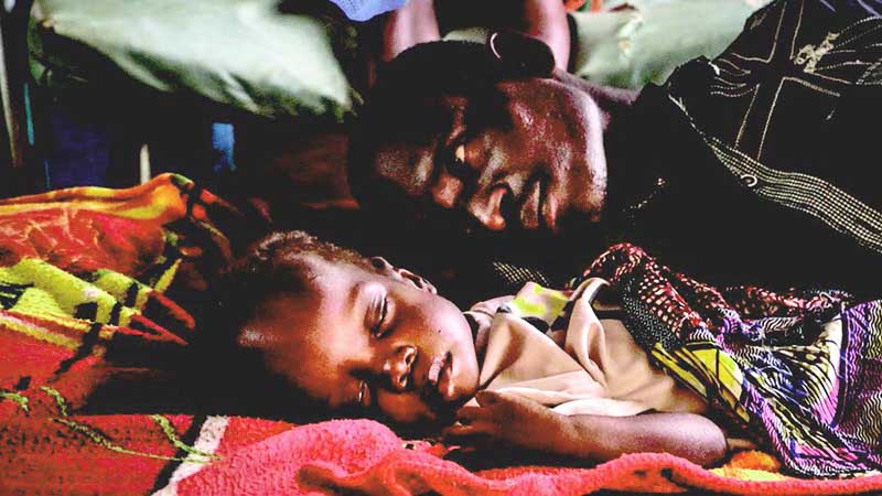 sick child in Central African Republic lying on a blanket with his father looking on