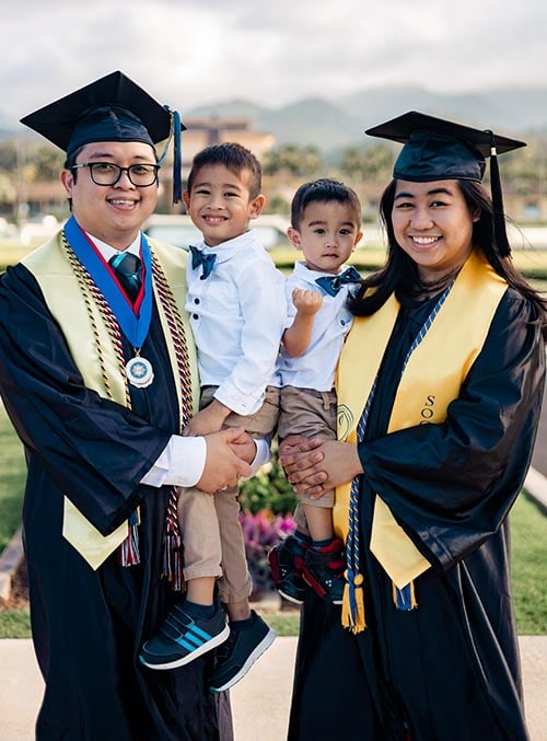 Husband and wife in graduation robes with their two young children