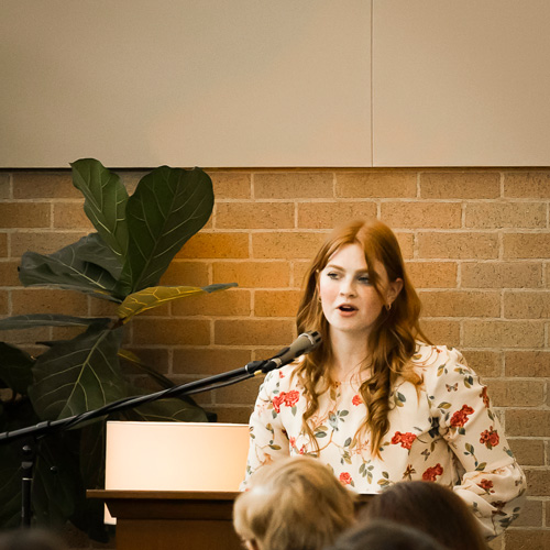 Headshot of young woman BYU student speaking into a microphone with red brick background behind her