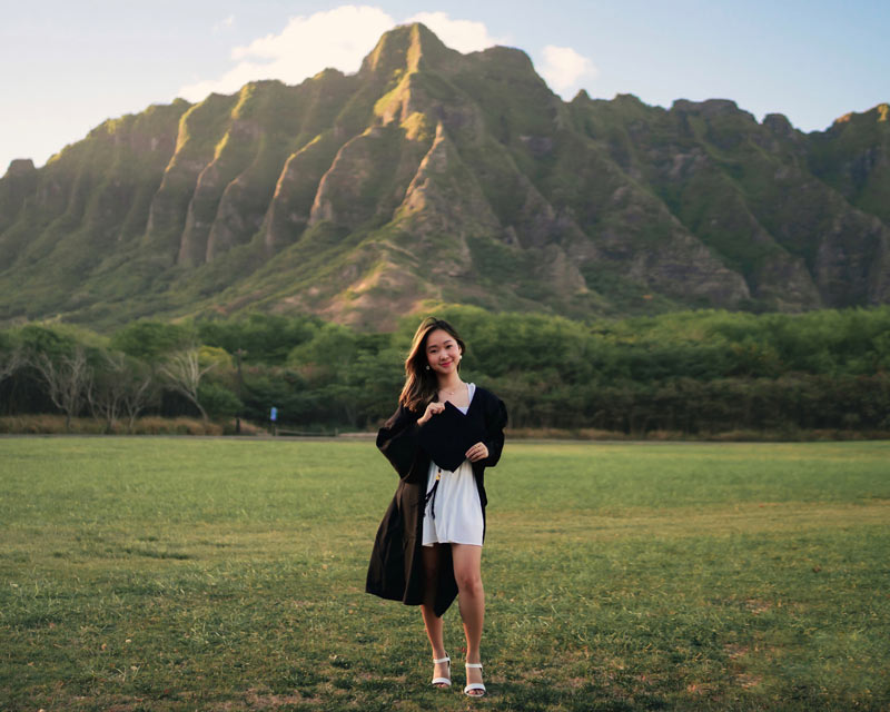 Crystal Tania in her graduation robe and holding her graduation cap, standing in front of a green mountain in Hawaii