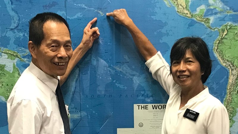 An elderly Chinese man and woman standing in front of a map pointing at Hawaii.