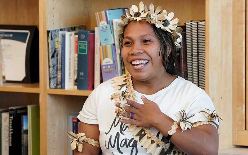 A Micronesian women, wearing flowers on her arms, over her shoulder, and in her hair, with one hand over her heart as to indicate gratitude.