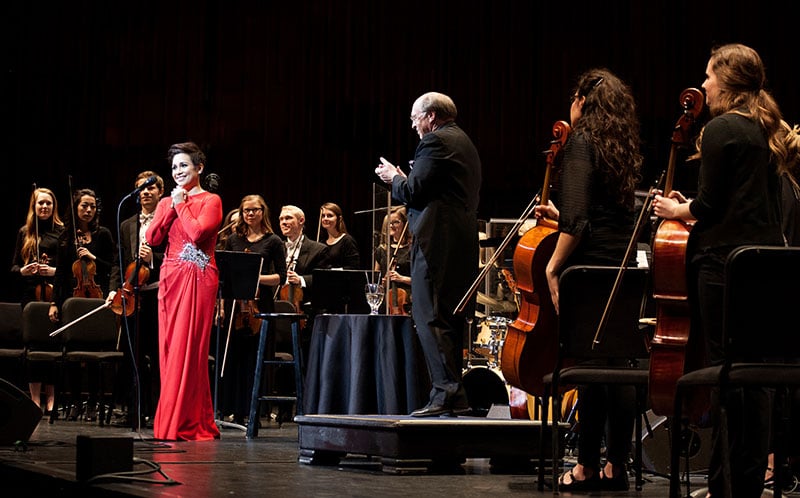 Female performer in red dress with orchestra and director standing during applause. 
