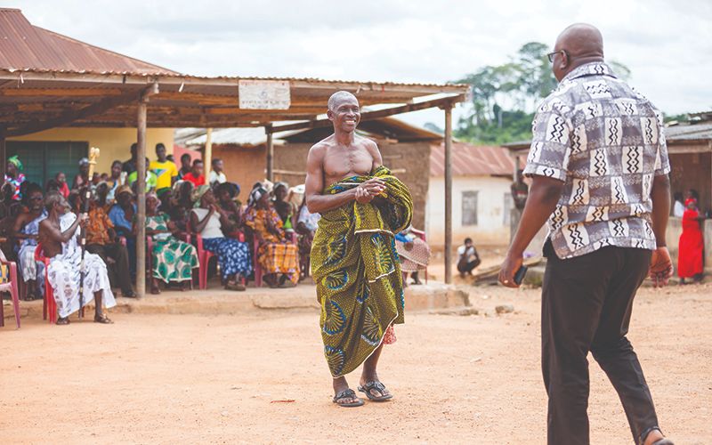 Photo taken in a village in Africa. Two men walk toward each other on a dirt road while a crowd of men, women, and children sit outside under the shade of a roof.