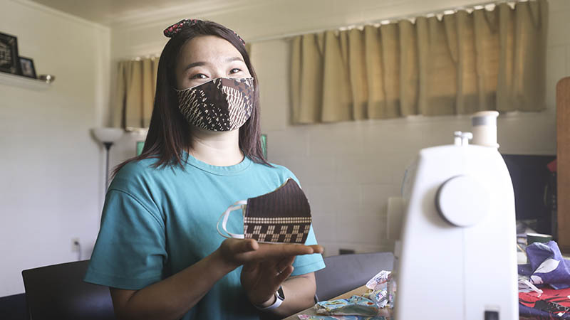 Asian woman sitting next to a sewing machine wearing and holding face masks