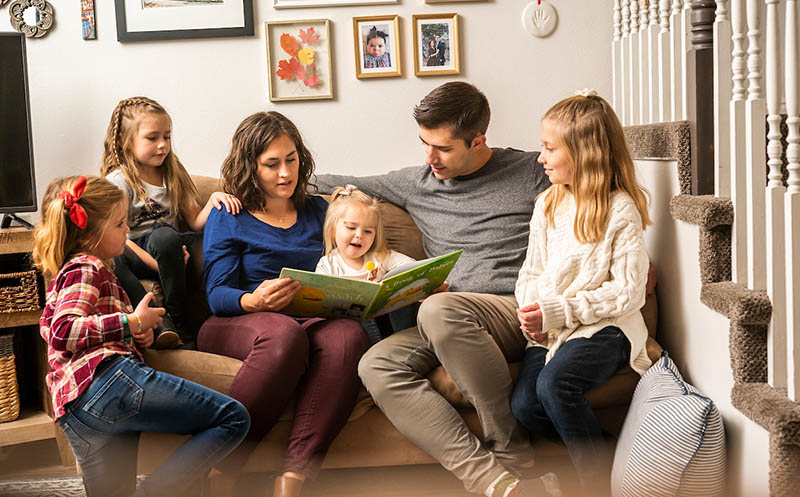 A mother and father sitting with their four young daughters and reading to the youngest, a toddler.