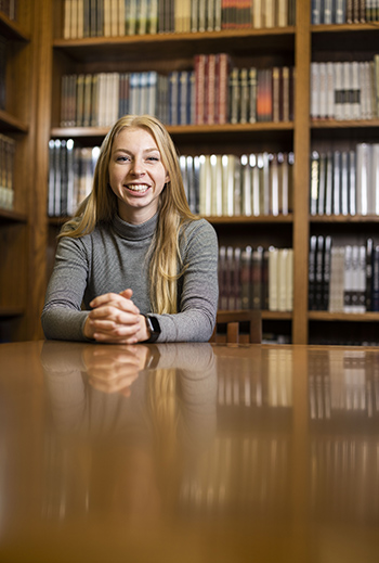 Smiling female seated at table in front of bookcase.